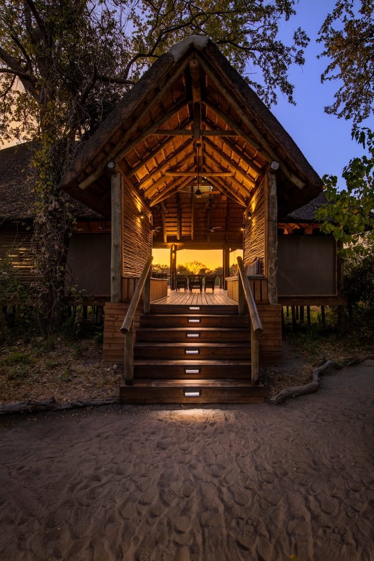 The lantern-lit entrance to the main area at Camp Khwai at dusk, with the sunset showing through to the deck beyond.