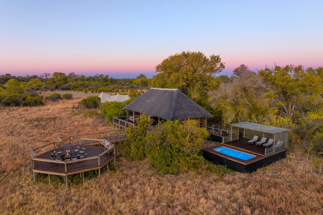 Camp Khwai from the air at dusk, showing the fire deck, thatched main area and pool set in open bush.