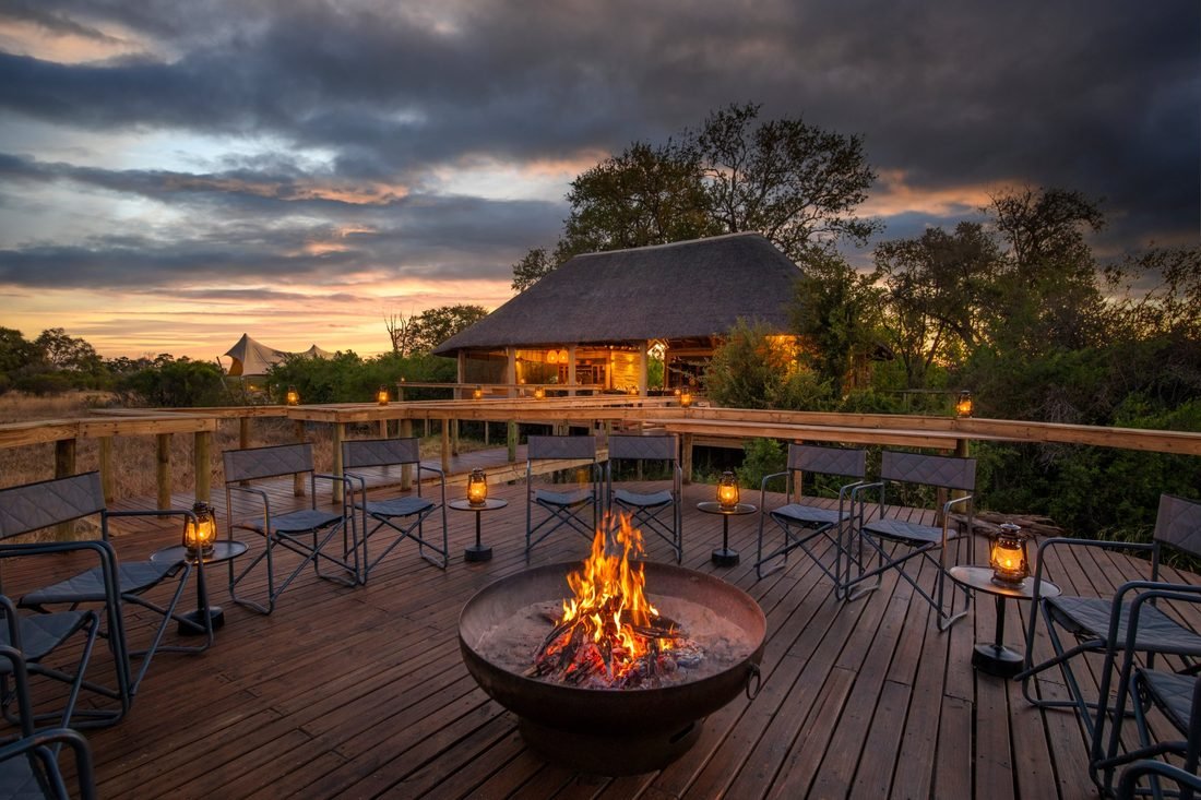 The fire pit burning on the deck at Camp Khwai at sunset, chairs set out facing the thatched main area.